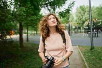 Фотограф-женщина с современной камерой стоит в зеленом парке и смотрит вверх — стоковое фото