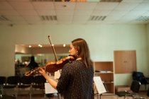 Зворотній вигляд зосередженої професійної жінки-музикантки, яка грає на акустичній скрипці та дивиться на музичний аркуш під час репетиції в студії — стокове фото