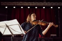 Músico feminino focado tocando violino com os olhos fechados perto do estande com partituras em luzes brilhantes na sala de concertos — Fotografia de Stock