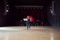 Músico feminino focado tocando violino perto do estande com partitura em luzes brilhantes na sala de concertos — Fotografia de Stock