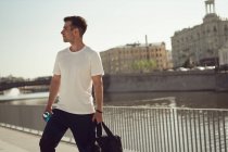 Contenuto atleta di sesso maschile con borsa sportiva e bottiglia d'acqua in piedi su argine in città in estate — Foto stock