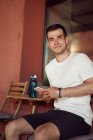 Smiling athlète masculin assis dans un café de rue avec une bouteille d'eau et en utilisant un téléphone mobile après l'entraînement en ville en regardant la caméra — Photo de stock