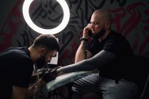Serious male tattoo master using tattoo machine while making tattoo on hand of guy with phone in light tattoo studio — Stock Photo