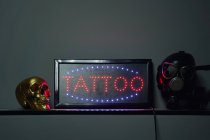 Светящаяся вывеска с татуировкой красного слова вокруг синих неоновых огней на полке рядом с золотым поддельным черепом и респиратором рядом с черной стеной в студии легких татуировок — стоковое фото