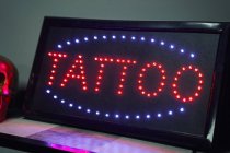 Glowing signboard with red word tattoo around blue neon lights on shelf next to golden fake skull and respirator near black wall in light tattoo studio — Stock Photo