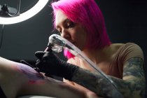 Серйозний майстер татуювання з рожевим волоссям в рукавичках з використанням професійного тату-машини при виготовленні татуювання на плечах клієнта в сучасному салоні татуювання. — стокове фото