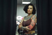 Mulher adulta séria em roupas casuais com tatuagens em pé no salão de tatuagem leve enquanto olha para a câmera com as mãos cruzadas — Fotografia de Stock
