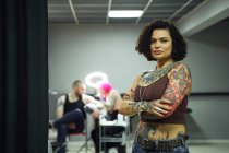 Серйозна доросла жінка в повсякденному одязі з татуюваннями, що стоять у світлому татуюванні салону, дивлячись на камеру з схрещеними руками — стокове фото