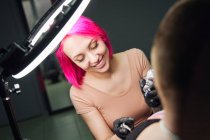 Maître de tatouage avec des cheveux roses dans des gants en utilisant une machine à tatouer professionnelle tout en faisant le tatouage sur l'épaule du client dans le salon de tatouage moderne — Photo de stock