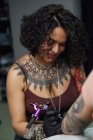 Maître de tatouage dans des gants en utilisant une machine à tatouer professionnelle tout en peignant le tatouage sur le bras de la femme dans un studio de tatouage moderne — Photo de stock