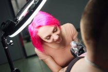 Mestre de tatuagem com cabelo rosa em luvas usando máquina de tatuagem profissional ao fazer tatuagem no ombro do cliente no salão de tatuagem moderno — Fotografia de Stock