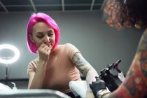 Mestre de tatuagem em luvas usando máquina de tatuagem profissional ao pintar tatuagem no braço da mulher no estúdio de tatuagem moderno — Fotografia de Stock