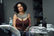 Pensive adult woman in casual clothes with tattoos sitting in light tattoo salon while looking at camera — Stock Photo