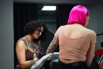 Maestro del tatuaje en guantes usando la máquina profesional del tatuaje mientras que pinta el tatuaje en el brazo de la mujer en el estudio moderno del tatuaje - foto de stock