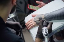 Концентрований чоловічий татуювач в рукавичках робить татуювання на руці клієнта, використовуючи професійну татуювальну машину в сучасній татуювальній студії — стокове фото