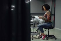 Mujer adulta seria en ropa casual con tatuajes sentados en el salón de tatuajes de luz mientras mira hacia abajo - foto de stock