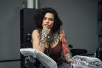 Серйозна доросла жінка в повсякденному одязі з татуюваннями, сидячи в легкому татуюванні салоні, дивлячись на камеру — стокове фото