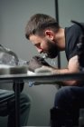 Tatuaje masculino concentrado en guantes que hacen el tatuaje en la mano del cliente mientras que usa la máquina profesional del tatuaje en estudio moderno del tatuaje - foto de stock