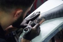 Tatoueur masculin concentré dans des gants faisant le tatouage sur la main du client tout en utilisant une machine à tatouer professionnelle dans un studio de tatouage moderne — Photo de stock