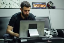 Удумливий дорослий хлопець у повсякденному вбранні з татуюваннями на комп'ютері в яскравій студії татуювання — стокове фото