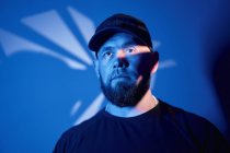 Selbstbewusster bärtiger erwachsener Mann in lässiger Kleidung und Mütze, der in einem dunklen Raum mit blauem Neonlicht und Schatten in die Kamera blickt — Stockfoto