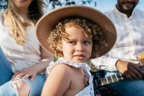 Deliziosa bambina con cappello che guarda lontano seduta con la famiglia multirazziale godendo pic-nic insieme mentre suonano la chitarra in natura — Foto stock