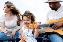 Famille multiraciale ravie de pique-niquer ensemble tout en mangeant des collations et en jouant de la guitare dans la nature — Photo de stock