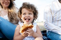 Весела маленька дівчинка їсть тістечко, дивлячись в сторону, сидячи з багаторасовою сім'єю, насолоджуючись пікніком разом у природі — стокове фото