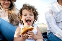 Веселая маленькая девочка ест тесто, смотрит в камеру, сидя с многорасовой семьей, наслаждаясь пикником на природе. — стоковое фото
