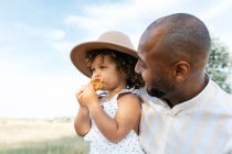 Positivo amorevole padre afroamericano abbracciando figlia adorabile mangiare panino gustoso nel campo estivo — Foto stock