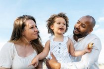 Baixo ângulo de família multiétnica feliz com filhinha fofa curtindo piquenique de verão na natureza — Fotografia de Stock