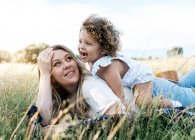 Vista lateral de la alegre madre y la niña de pelo rizado acostados juntos en la manta en el prado y disfrutando del día de verano - foto de stock