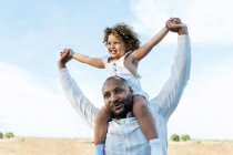 Alegre pai afro-americano com filhinha fofa nos ombros jogando em campo no verão e se divertindo olhando para longe — Fotografia de Stock