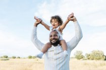 Cheerful African American father with cute little daughter on shoulders playing in field in summer and having fun looking away — Stock Photo
