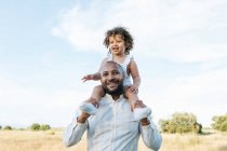 Alegre padre afroamericano con linda hija sobre hombros jugando en el campo en verano y divirtiéndose mirando hacia otro lado - foto de stock