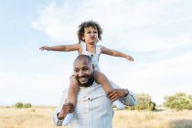 Fröhlicher afroamerikanischer Vater mit süßer kleiner Tochter auf den Schultern, die im Sommer auf dem Feld spielt und Spaß beim Wegschauen hat — Stockfoto
