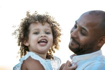 Отец афроамериканец держит милую кудрявую афроамериканскую дочь на фоне закатного неба в природе, смотрящую в камеру — стоковое фото