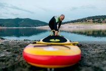 Мужчина-серфер в гидрокостюме накачивает доску SUP, стоя на берегу моря и готовится к серфингу — стоковое фото