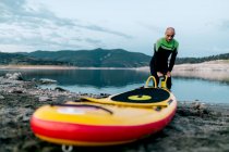 Мужчина-серфер в гидрокостюме накачивает доску SUP, стоя на берегу моря и готовится к серфингу — стоковое фото