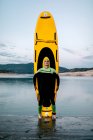 Спокійний чоловічий серфер у гідрокостюмі, що стоїть очі закриті жовтим дошкою СУП на пляжі біля моря — стокове фото