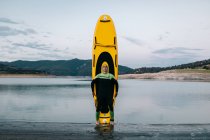 Calm male surfer in wetsuit standing with eyes closed with yellow SUP board on beach near sea — Stock Photo