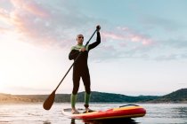 Von unten fokussierter männlicher Surfer, der bei Sonnenuntergang im Sommer auf dem SUP-Board auf ruhiger See schwimmt — Stockfoto