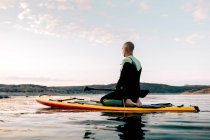 Side view of tranquil male surfer sitting in Thunderbolt pose on paddleboard and mediating while practicing yoga in sea in evening — Stock Photo