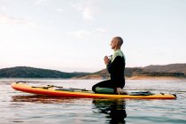 Вид сбоку спокойного серфера-мужчины, сидящего в Thunderball позировать с Намасте руками на поддоне и посредничать, практикуя йогу в море по вечерам — стоковое фото