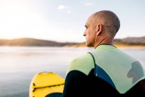 Вид сзади на взрослого мужчину в гидрокостюме, стоящего на коленях на гребной доске на спокойном озере — стоковое фото