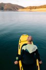 De cima visão traseira do macho adulto em wetsuit ajoelhado na tábua de remo na superfície calma da água do lago — Fotografia de Stock