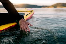 Cultive surfista masculino irreconhecível sentado na placa SUP e tocando a água do mar ao pôr do sol — Fotografia de Stock