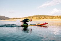 Full body side view of adult male in wetsuit kneeling on paddle board and paddling with arms on calm lake water surface — Stock Photo