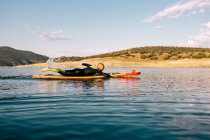 Босоногий мужчина в гидрокостюме лежит на доске и плавает на поверхности озера во время занятий водными видами спорта в летний день — стоковое фото