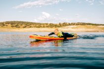 Side view of barefoot male in wetsuit lying on paddle board and swimming on lake surface while practicing water sport in summer day — Stock Photo